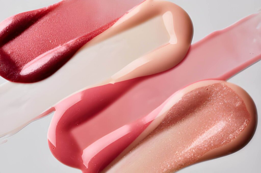 Swatches of various lip products in shades of pink, coral, and nude, showcasing different finishes and textures on a white background.