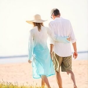 A woman in a sun hat and a man in shorts walk arm in arm on a sunny beach. They appear relaxed, with a gentle sea and sand in the background.