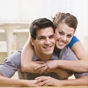 A smiling man and woman embrace indoors, conveying a joyful and affectionate mood. The woman is playfully hugging the man from behind.