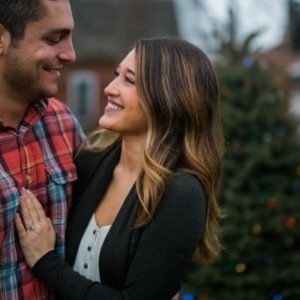 A couple embraces and smiles at each other warmly, both wearing winter clothes. A blurred Christmas tree with colorful lights stands in the background.