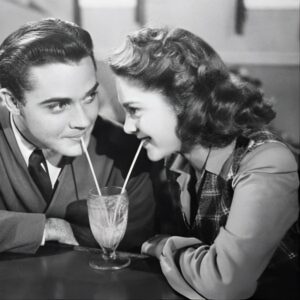 A black and white photo of a smiling couple sharing a milkshake with two straws across a table. They appear to be in a retro diner, creating a nostalgic and romantic atmosphere.