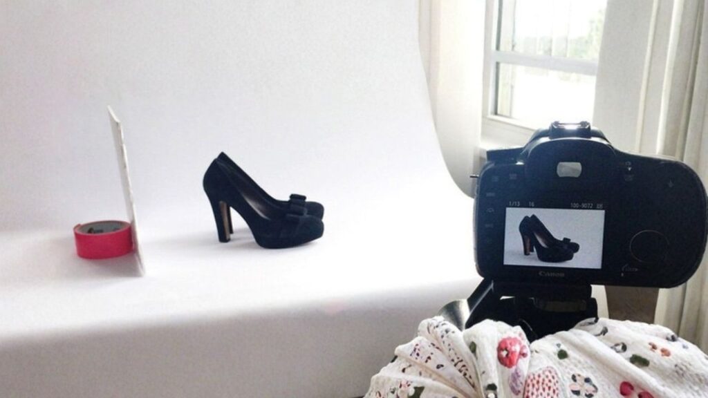 A black high-heeled shoe is displayed on a white backdrop, with a camera capturing its image. Nearby, a red roll of tape and a window with soft natural light.