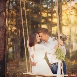 A couple embraces on a wooden swing in a sunlit forest. The man leans in to kiss the woman's forehead, conveying warmth and romance amid autumn trees.