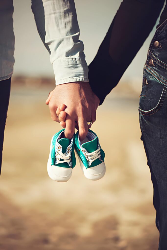A couple holds hands, with one hand grasping a pair of small turquoise children's shoes, set against a sandy background.