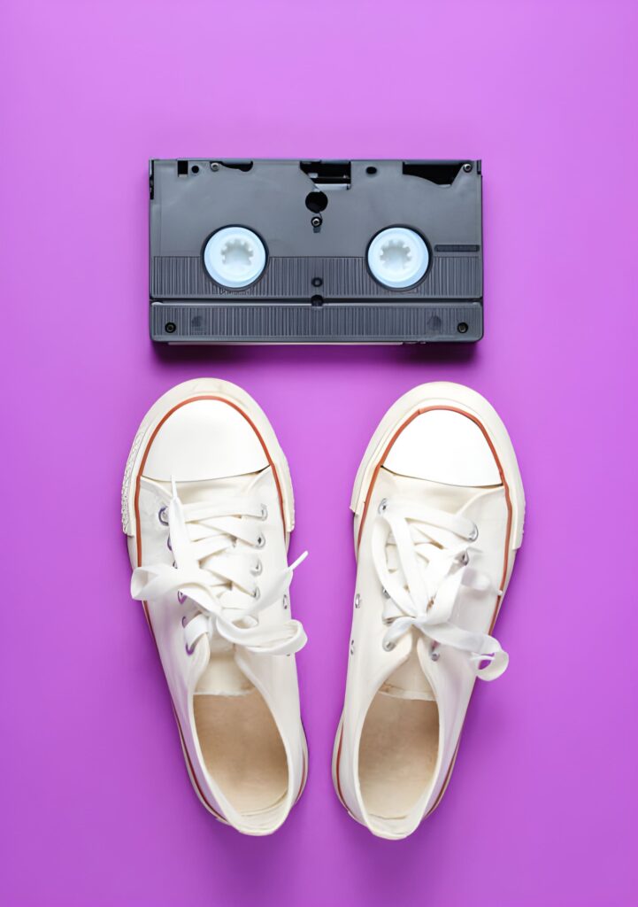 A pair of white sneakers rests on a vibrant purple background, above a black VHS tape with visible reels.
