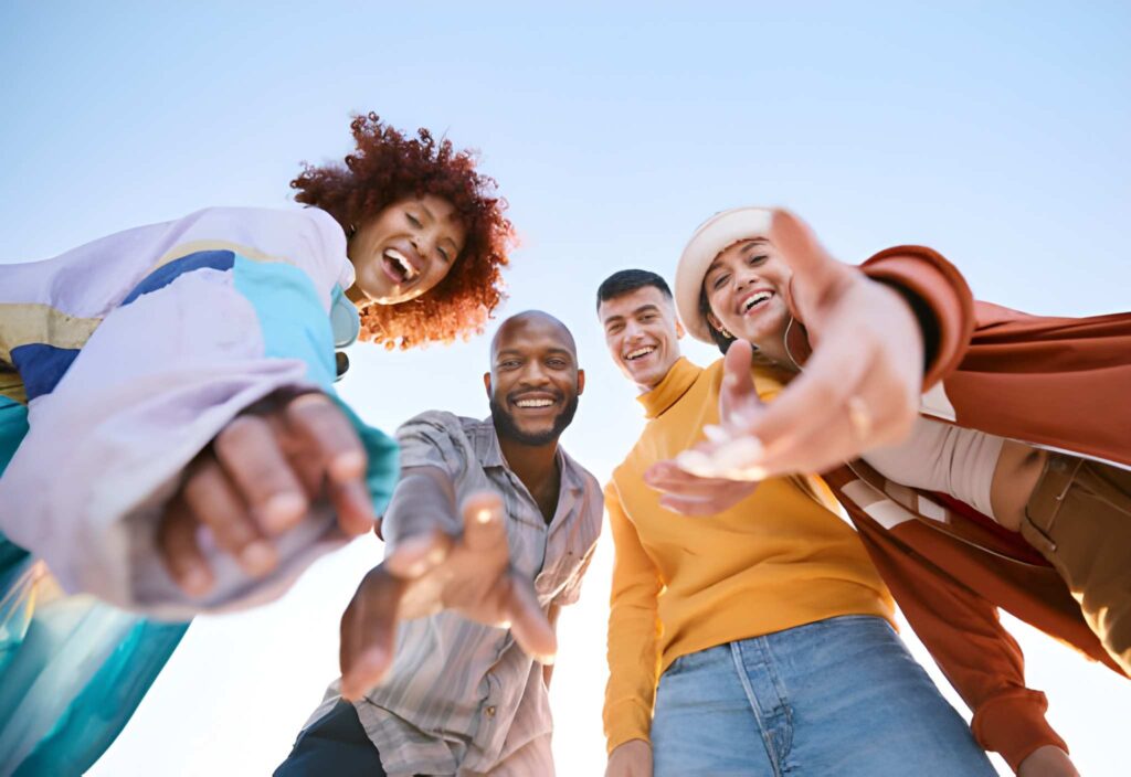 Low-angle view of a diverse group of young people smiling and reaching toward the camera outdoors