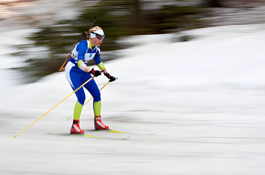 Dynamic pan-shot photograph of a winter sports athlete skiing at high speed while carrying a rifle, captured with motion blur to emphasize movement and athletic performance.