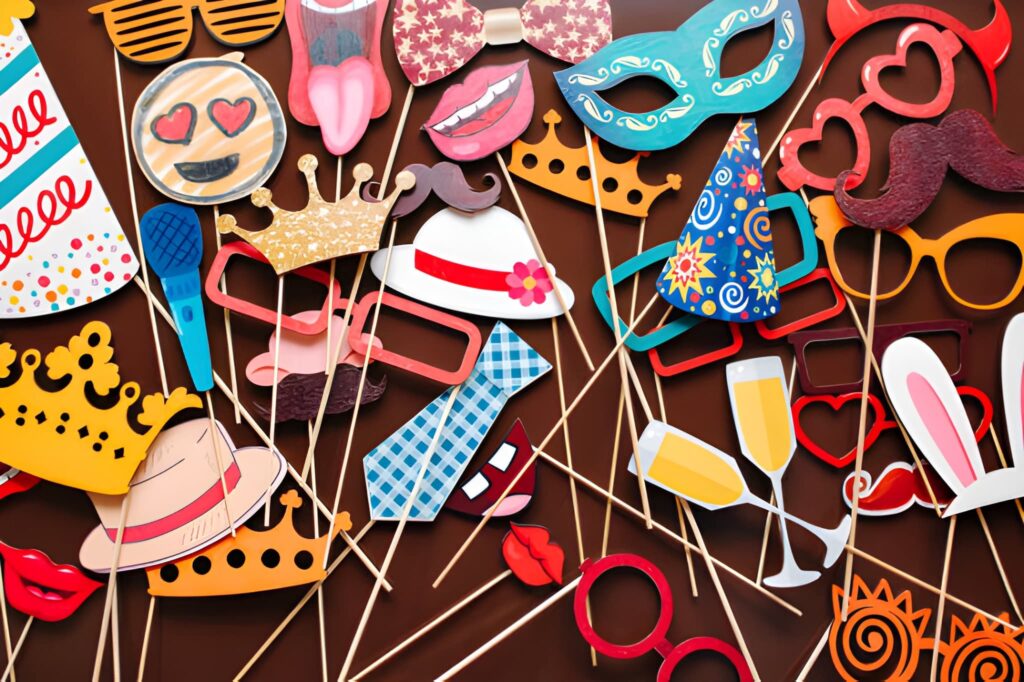 A flat lay arrangement of colorful and creative photo booth props, including glasses, masks, hats, crowns, lips, mustaches, and party-themed accessories on wooden sticks.