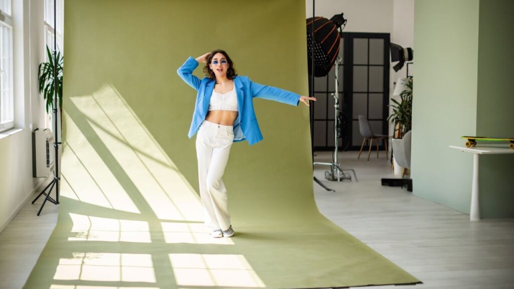 A model poses playfully in a bright studio, wearing a blue jacket and white pants, against a green backdrop with natural light.