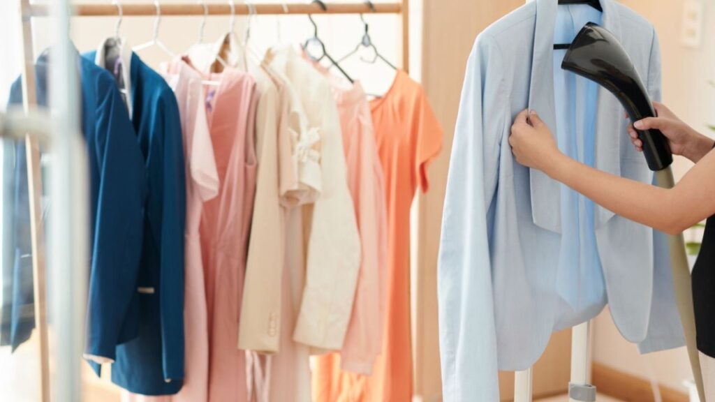 A person uses a handheld garment steamer on a light blue jacket hanging on a rack alongside several other colorful, neatly hung shirts and dresses.