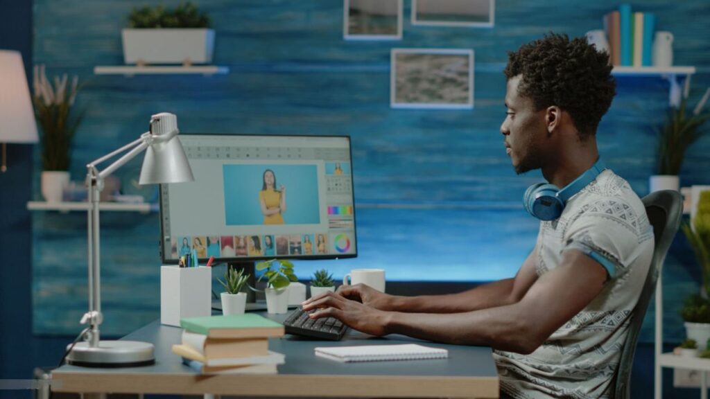 Young Black man editing a photograph on a desktop computer at a modern desk illuminated by a silver task lamp.