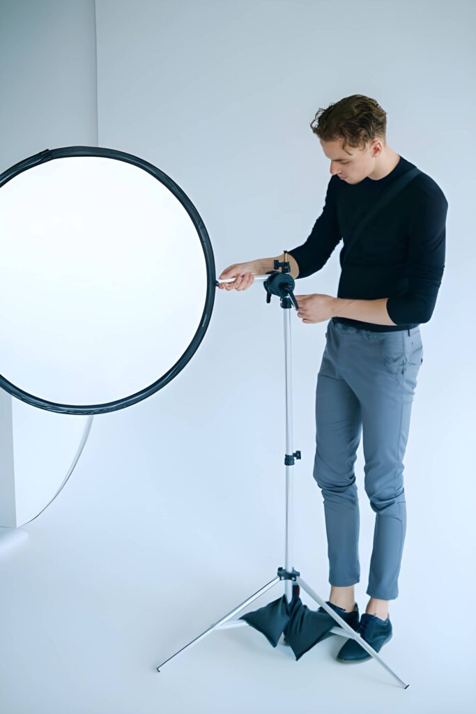 A person stands near a round reflector on a tripod, adjusting its height in a bright, minimalistic studio.