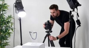 A man uses a camera on the tripod to photograph a necklace on a table and a lighting setup and a tree are beside it.
