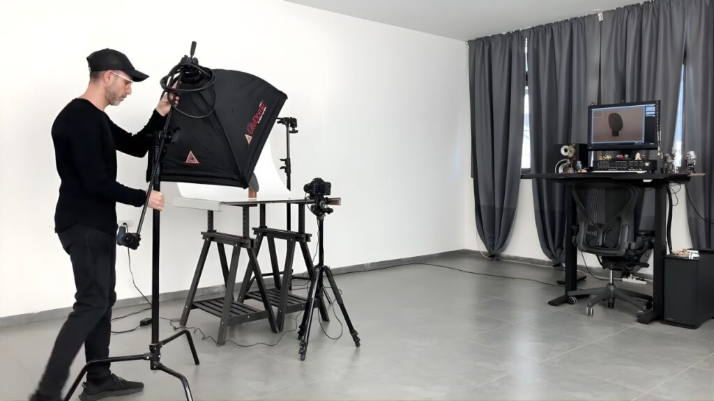 A photographer in a black outfit adjusts studio lighting for a product shoot. Nearby, a camera is set up on a tripod, and a computer displays images. The setting conveys professionalism and focus.