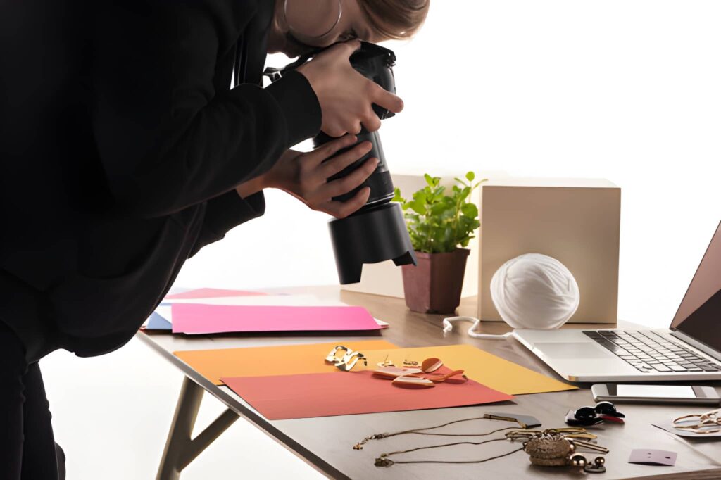 Person photographing jewelry on a desk with colorful paper. A laptop, potted plant, and white object are nearby. The scene feels creative and focused.