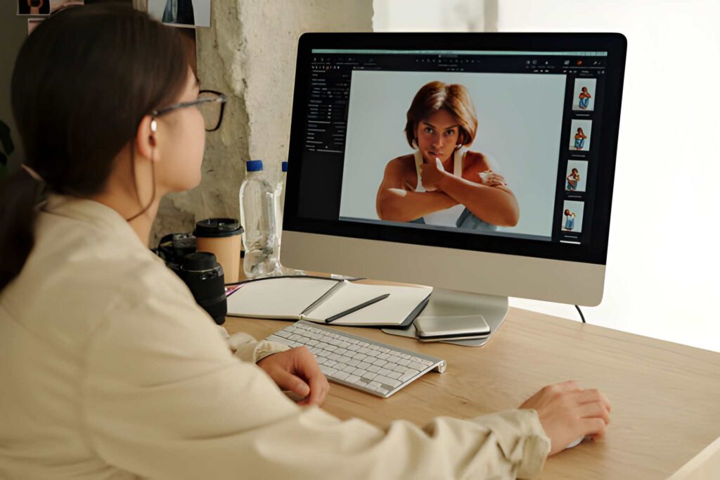 A person sits at a computer desk, editing photos on a large monitor with images of a model displayed on the screen.