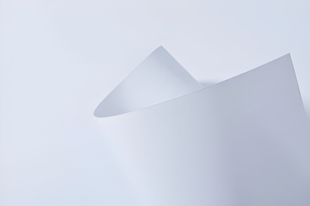 A close-up of a curved, white sheet of paper against a light gray background, creating a minimalist and abstract composition.