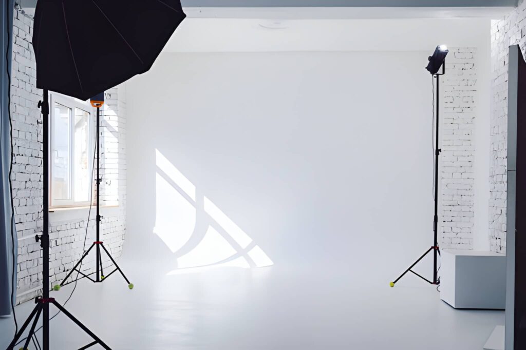Photography studio with a white backdrop, brick walls, sunlight streaming through a window, and two black light stands. Minimalistic and bright.