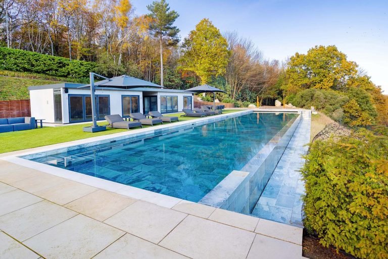 A luxurious outdoor pool with clear blue water, surrounded by sun loungers and a modern house with large glass windows, set against a backdrop of autumn-colored trees.