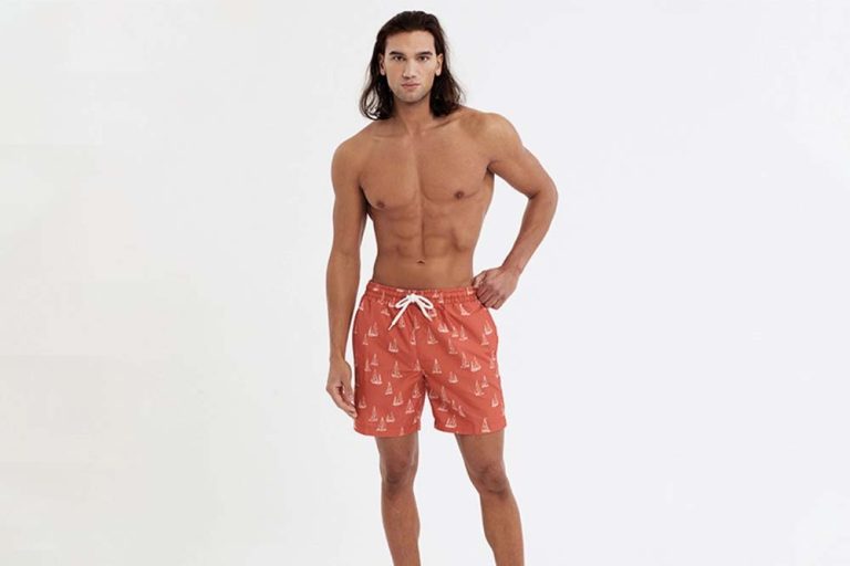 A male model with long hair stands confidently wearing orange swim shorts with a white sailboat pattern, against a plain white background before alpha channel masking.