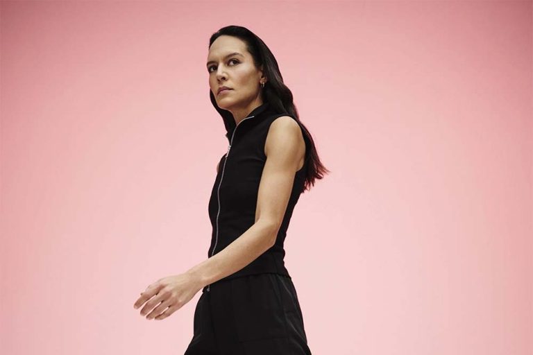 A woman in a black sleeveless outfit enhanced with color masking, walks confidently against a soft pink background. She appears focused, with her hair tied back.