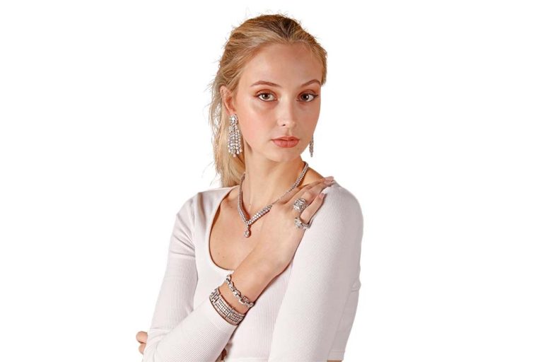 Young woman with blonde hair, wearing a white top and multiple pieces of jewelry, posing with her hands crossed against a white background after image masking.