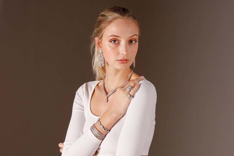 Young woman with blonde hair, wearing a white top and multiple pieces of jewelry, posing with her hands crossed against a plain background.