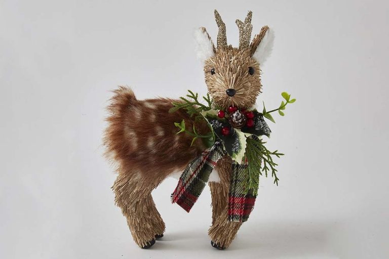 A decorative figurine of a reindeer crafted from twigs and faux fur, adorned with a small plaid scarf and a sprig of holly berries on a white background.