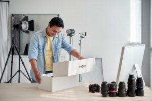 A person wearing a blue jacket and yellow shirt opens a white box on a table. Behind them are professional studio lights, a camera, and several camera lenses. A computer monitor and more camera equipment are presented.