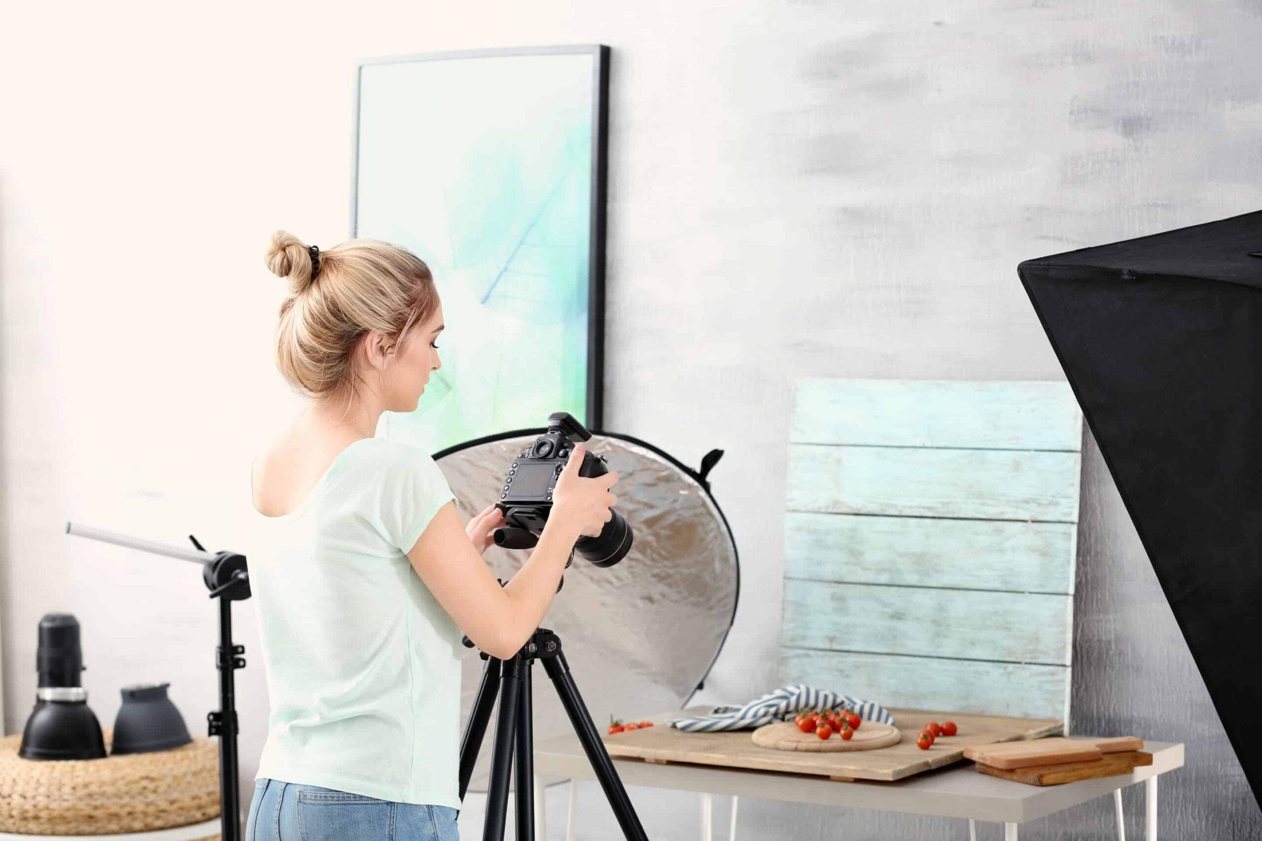 A woman is fixing camera in the DIY product photography setup. This picture includes lighting, reflector, backdrop and objects are placed on the table, and a tripod to prevent unwanted shakings.