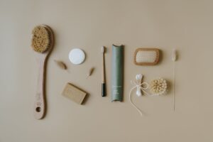A flat lay of eco-friendly personal care items arranged neatly on a beige surface. The items include a wooden body brush, soap bar, reusable cotton pads, a bamboo toothbrush, floss, a scrub sponge, and cotton.