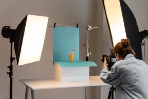 A photographer is capturing an image of a tropical drink with a small umbrella on a white table. The drink is set against a blue backdrop, with professional lighting equipment on both sides.