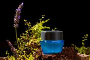 It is showing a blue box of beauty product on a clay surface, small plants, flower are around it are used as props to enhance the atmosphere. 