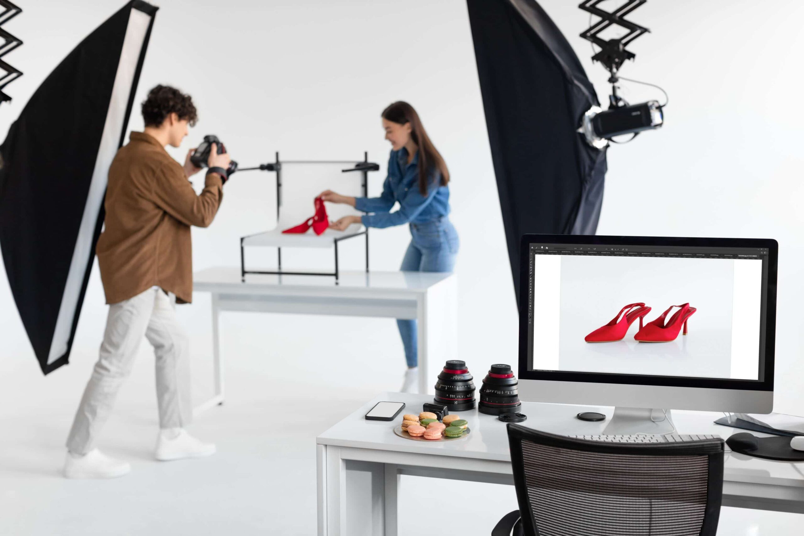Two models are showing product photography ideas, capturing a pair of red shoe. The picture is showing a desktop computer displaying the red shoe, two camera lens, and a plate of biscuits on the table. The picture also includes two lighting setup and a chair in front of the table.