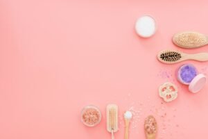 This is a flat lay product photography on a plain pink surface. In this composition objects are presented on a C-shape. The objects include three combs, three pots of organic product and two spoons carrying the same items. 