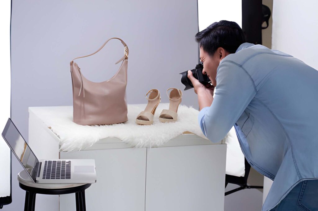 A person photographs a beige handbag and matching high-heeled sandals on a fluffy surface, with a laptop nearby for reference.