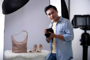 Ecommerce product photography setup with a person holding a camera, capturing a pink handbag and high-heeled sandals on a white backdrop.