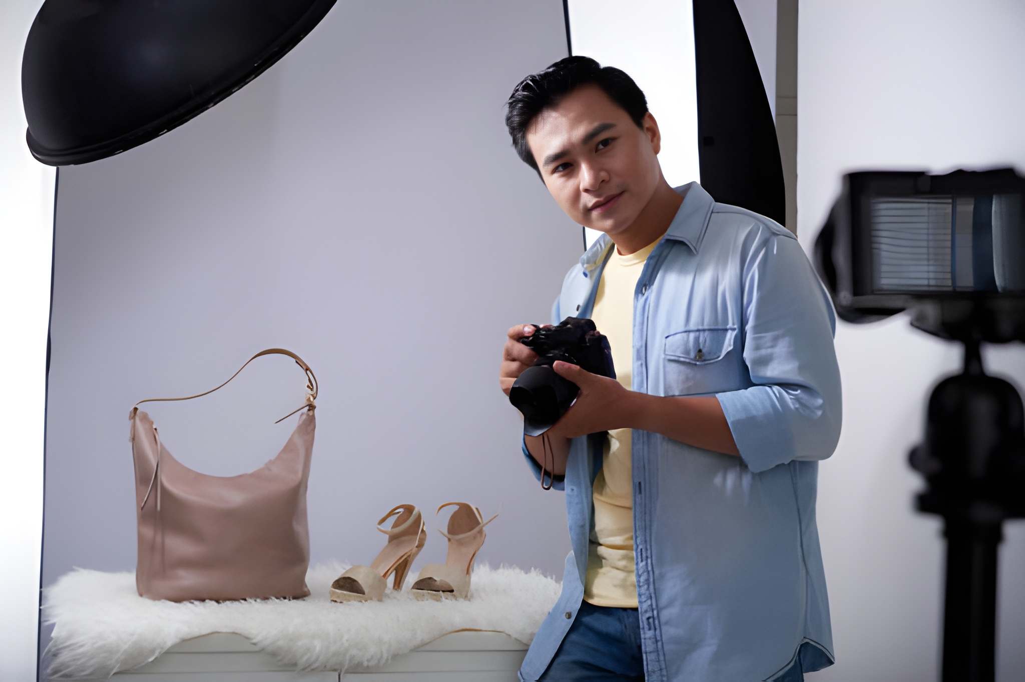 Ecommerce product photography setup with a person holding a camera, capturing a pink handbag and high-heeled sandals on a white backdrop.