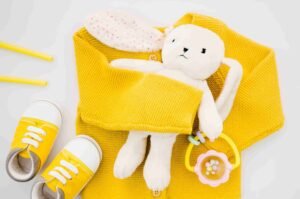 A plush rabbit toy with a yellow knit sweater, white and yellow baby shoes, and a rattle, representing a lifestyle product photography.