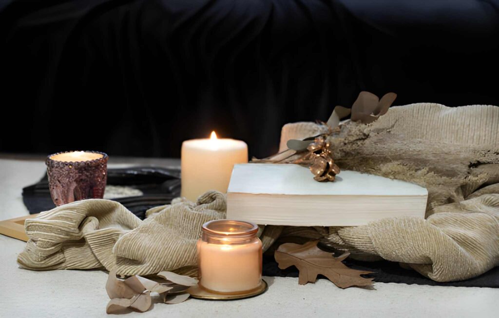 A cozy arrangement featuring candles, a closed book with a decorative piece, and soft fabric, surrounded by autumn leaves.