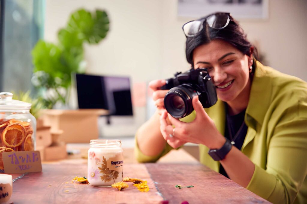 A person is capturing photos with a camera in a bright, modern workspace decorated with plants and various candle jars on a wooden table.