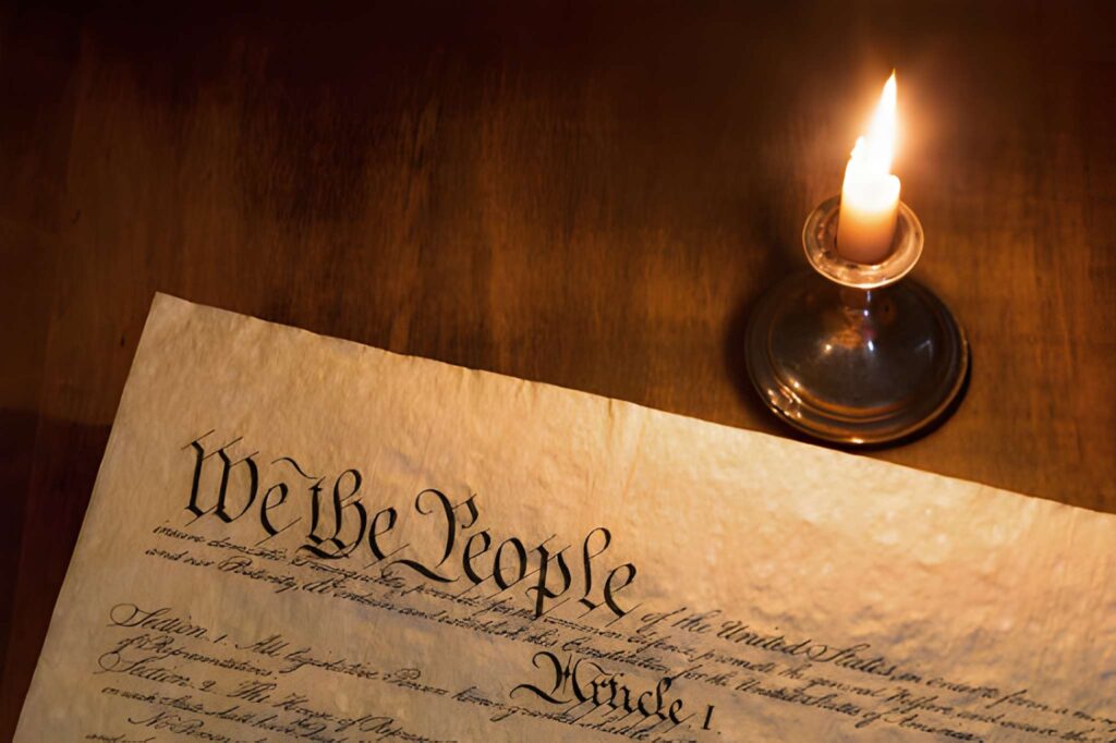 A candle illuminates a historic parchment titled "We the People," resting on a wooden table. The scene evokes a sense of history and solemnity.