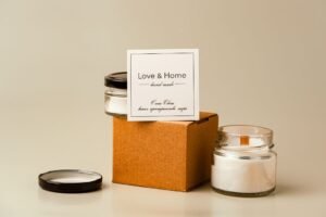 Handmade candle products with a "Love & Home" label on a cardboard box, two candles against a beige background.