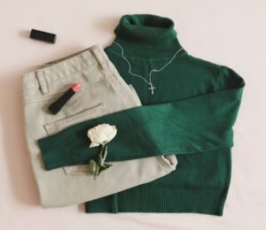 A green turtleneck sweater paired with green pants, accented by a white rose, lipstick, and a cross pendant on a neutral background.