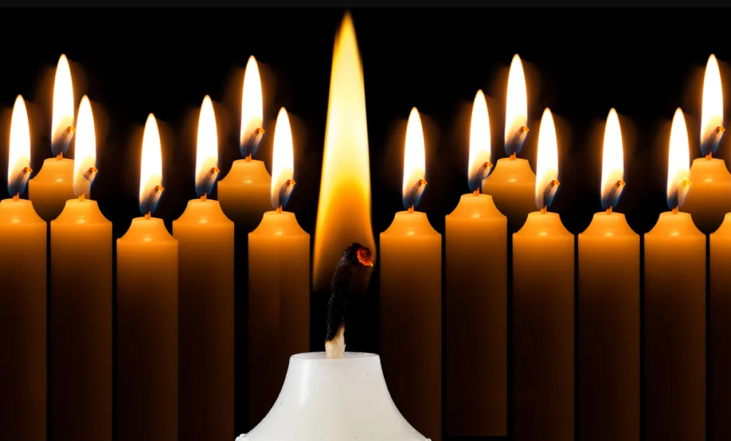 A single lit candle in the foreground is surrounded by numerous blurred candles in the background against a dark backdrop, creating a warm, serene glow.