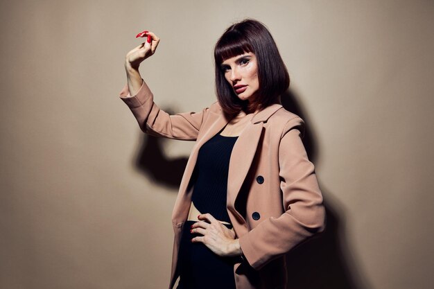 A person in a stylish beige coat poses confidently against a neutral background, holding a red object in one hand.