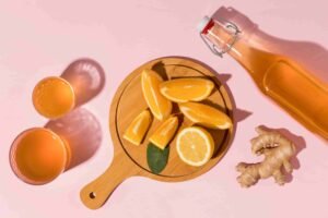 Sliced oranges on a cutting board with ginger, a bottle, and glasses of juice on a pink background, presenting a flat-lay style product photography.