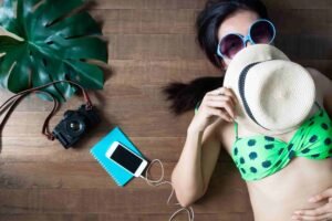 A woman wearing a green polka dot bikini and blue sunglasses covers her face with a hat. Next to her are a leaf, a camera, a notebook, and a phone with earphones attached, all on a wooden floor, representing lifestyle product photography.