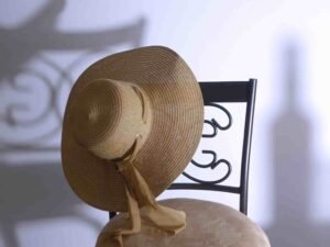 A wide-brimmed straw hat with ribbons is elegantly placed on a decorative metal chair, its shadow artfully cast on the wall, capturing the essence of lifestyle product photography.