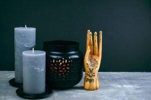 Two grey candles, a black candle holder with cut-out patterns, and a wooden hand sculpture with a sun design are arranged on a surface against a dark background.